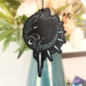 May include: Black and white wolf head illustration with a crescent moon and geometric patterns. The image is on a car air freshener.