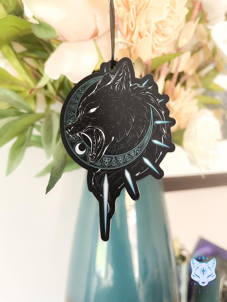 May include: Black and white wolf head illustration with a crescent moon and geometric patterns. The image is on a car air freshener.