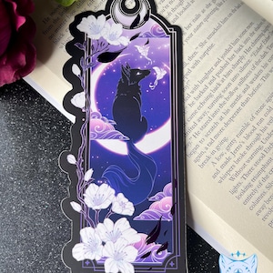 Può includere: Segnalibro decorativo con una silhouette di lupo nero su una luna crescente su uno sfondo viola stellato. Fiori di ciliegio bianchi e nuvole rosa incorniciano il disegno. Il segnalibro ha un bordo nero.