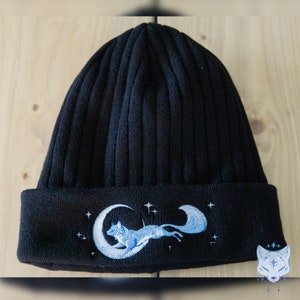 May include: Black knit beanie with a blue embroidered design of a fox running on a crescent moon with stars.