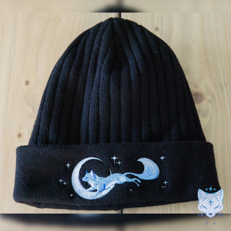 May include: Black knit beanie with a blue embroidered design of a fox running on a crescent moon with stars.