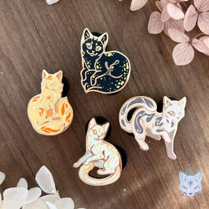 May include: Four enamel pins featuring different colored cats with gold outlines. The cats are in various poses, including curled up, sitting, and standing. The pins are on a wooden surface.