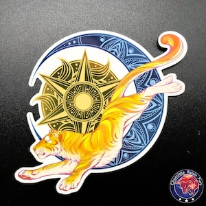 May include: A colorful sticker featuring a stylized orange and yellow tiger leaping through a crescent moon and sun design. The sun is a six-pointed star with a yellow center and the moon is a blue crescent with a mandala pattern.