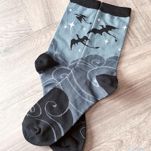 May include: A pair of crew socks with a dragon and star design. The socks have a light blue background with dark grey clouds and black dragon silhouettes. The toes and heels are black.