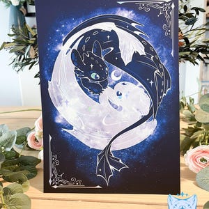 May include: A dark blue print depicting two dragons, one black and one white, within a glowing moon shape against a starry backdrop. The artwork has decorative corner elements and a celestial theme.