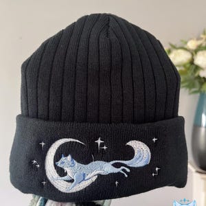 May include: Black knit beanie with a white embroidered design of a fox jumping over a crescent moon with stars.