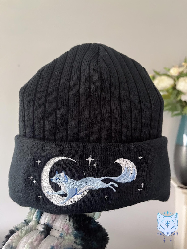 May include: Black knit beanie with a white embroidered design of a fox jumping over a crescent moon with stars.