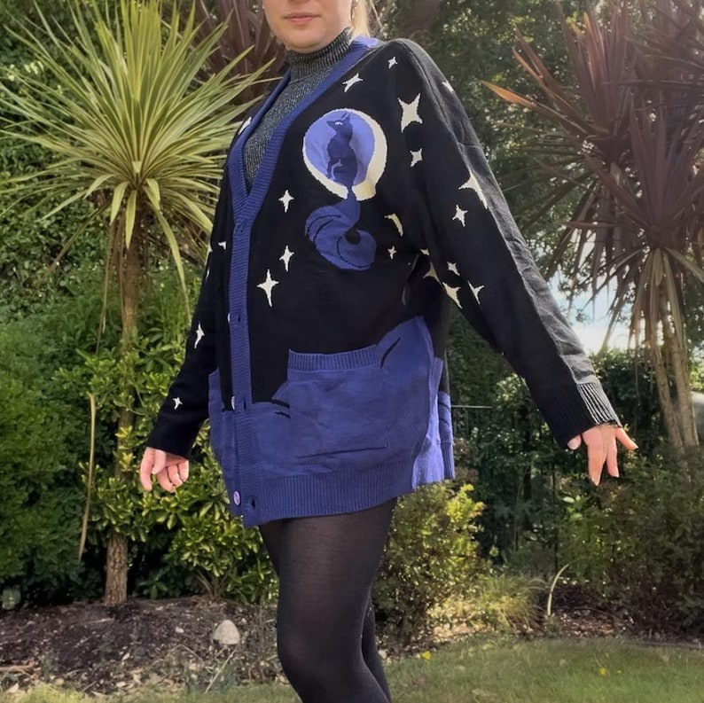 May include: Black cardigan sweater with a blue cat and moon design, white stars, and blue pockets. The sweater has a button-up front and ribbed cuffs and hem. A unique and stylish piece of clothing.