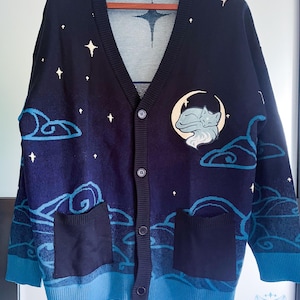 May include: Navy blue cardigan sweater with a celestial design. The sweater features a v-neck, button closure, and two front pockets. The design includes white stars, clouds, and a moon with a sleeping fox. The cuffs and bottom hem are a lighter shade of blue.