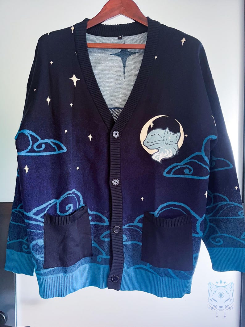 May include: Navy blue cardigan sweater with a celestial design. The sweater features a v-neck, button closure, and two front pockets. The design includes white stars, clouds, and a moon with a sleeping fox. The cuffs and bottom hem are a lighter shade of blue.