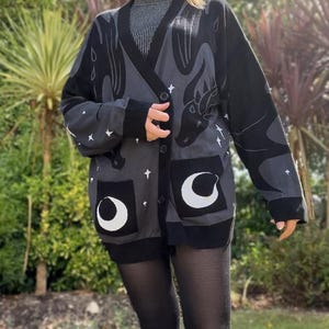 May include: A dark gray cardigan with a dragon design, white stars, and crescent moon pockets. The long-sleeved sweater has a black trim and buttons. The dragon design is on the back and sleeves.