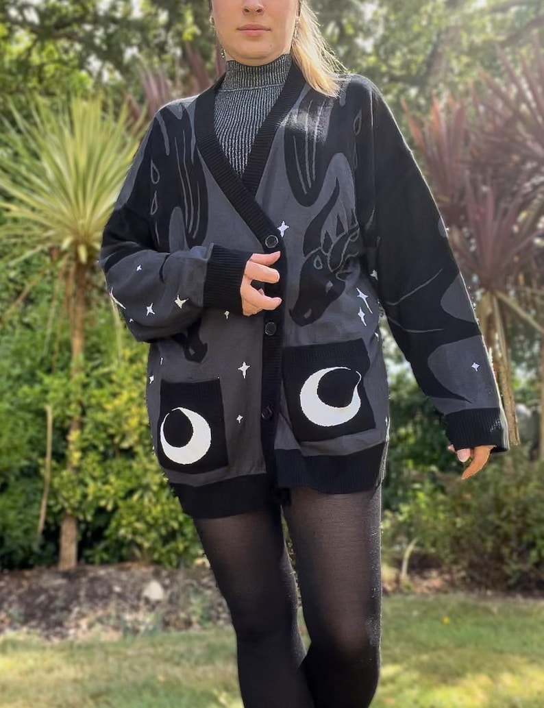 May include: A dark gray cardigan with a dragon design, white stars, and crescent moon pockets. The long-sleeved sweater has a black trim and buttons. The dragon design is on the back and sleeves.