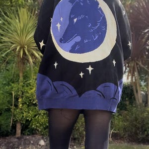 May include: Black knit sweater featuring a blue wolf silhouette inside a white crescent moon. The sweater has a black background with white stars and blue cloud-like details at the bottom. A unique and cozy piece.