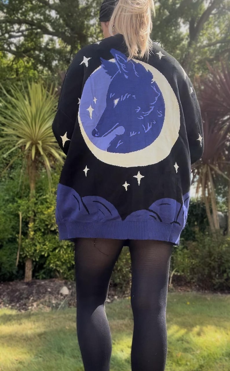 May include: Black knit sweater featuring a blue wolf silhouette inside a white crescent moon. The sweater has a black background with white stars and blue cloud-like details at the bottom. A unique and cozy piece.