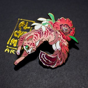 May include: A pink and red enamel pin of a cat with a flower in its hair. The cat is curled up in a ball with its tail wrapped around its body. The flower is a red poppy with a yellow center.