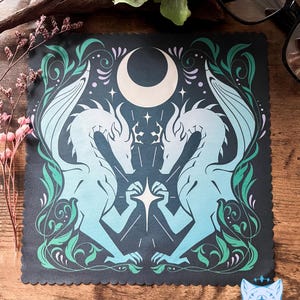May include: A dark blue square cloth featuring a celestial design with two stylized dragons facing each other. The dragons are light blue and have wings, with a crescent moon and stars above. Green and purple floral accents frame the image.
