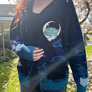 May include: Navy blue cardigan sweater with a celestial design. Features a sleeping fox on a crescent moon, clouds, and stars. The sweater has teal accents on the cuffs, pockets, and trim. A button-down front and two front pockets complete the design.
