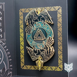 May include: Gold-toned metal pin with a black and white raven design, a blue and gold Valknut symbol, and a gold arrow. The pin is surrounded by a gold Celtic knot border.