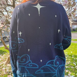 May include: Navy blue sweater with a celestial design. The sweater features a large star on the upper back and smaller stars scattered across the navy blue fabric. The bottom and cuffs are teal, and there are teal cloud designs.