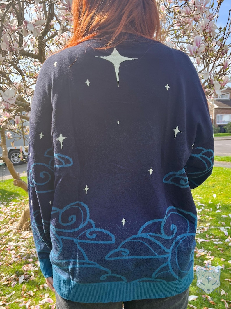May include: Navy blue sweater with a celestial design. The sweater features a large star on the upper back and smaller stars scattered across the navy blue fabric. The bottom and cuffs are teal, and there are teal cloud designs.
