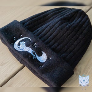May include: Black knit beanie with a white embroidered design of a fox curled around a crescent moon with stars.