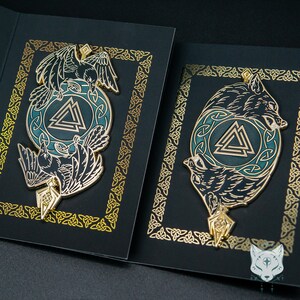 May include: Two black and gold enamel pins with a green and blue background. The pins feature a raven and a wolf, both with a triangular symbol in the center.