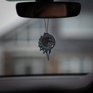 May include: A black and silver car air freshener with a wolf's head design. The wolf is snarling and has a blue eye. The air freshener is hanging from the rearview mirror of a car.