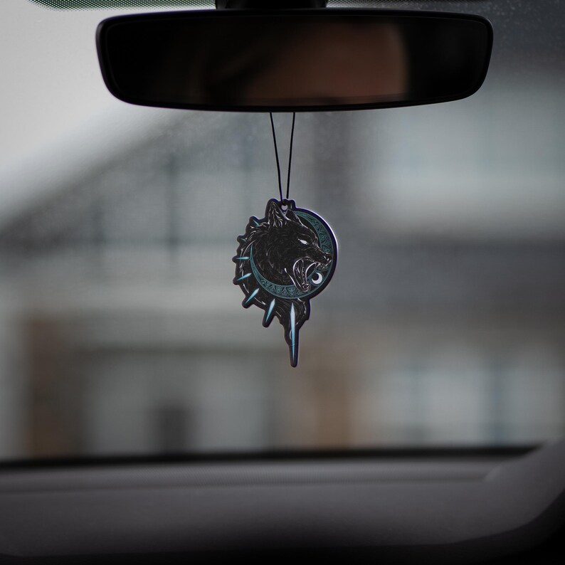May include: A black and silver car air freshener with a wolf's head design. The wolf is snarling and has a blue eye. The air freshener is hanging from the rearview mirror of a car.