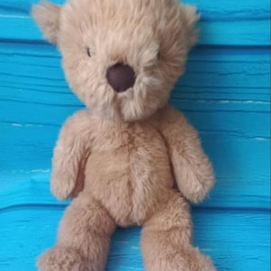 May include: A plush teddy bear with light tan, shaggy fur and a dark brown, round nose. The stuffed animal features small, rounded ears, a soft textured body, and limbs positioned in a seated posture, showcasing a simple and classic design.