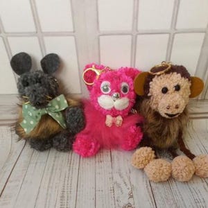 May include: Three plush animal keychains: a gray mouse with a green bow, a pink cat with a bow, and a brown monkey with pom-pom feet. The keychains are made of soft, fuzzy material and have gold-colored key rings.