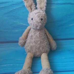 Op de afbeelding: Een zacht, grijs konijnenspeelgoed met lange oren, corduroy armen en benen en een pluizig lichaam. Het knuffeldier staat tegen een helderblauwe houten achtergrond.