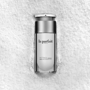 Puede incluir: Botella de sérum cosmético plateada y blanca con el texto "la parfait cosmetics" y "Retinol Serum Max Rejuvenation". La botella tiene un dispensador de bomba y está sobre un fondo blanco texturizado.