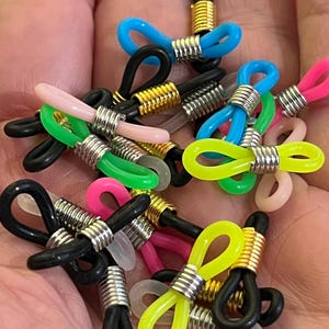 May include: A collection of colourful eyeglass chain holders with gold and silver metal accents. The holders are made of silicone and feature a bow shape.
