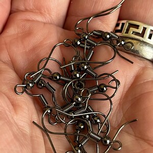 May include: A pile of dark grey earring hooks, each with a coiled spring and a small ball. The hooks are held in a hand, with a silver ring visible in the background. These earring findings are for jewellery making.