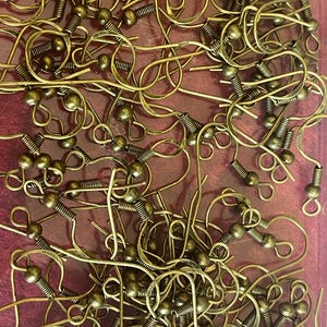 May include: A close-up of a collection of antique brass-toned earring hooks. The hooks feature coiled springs, curved wires, and small ball details. They are scattered across a reddish-purple surface, showing a variety of shapes and sizes.