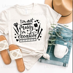 May include: A white t-shirt with black text that reads "I'm not messy I'm creative" with a ruler, scissors, and other craft supplies surrounding the text.