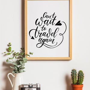 May include: A framed print with the text "Can't wait to travel again" in black calligraphy with a paper airplane graphic. The print is on a white background and is framed in a light wood frame. The print is hanging on a white wall.