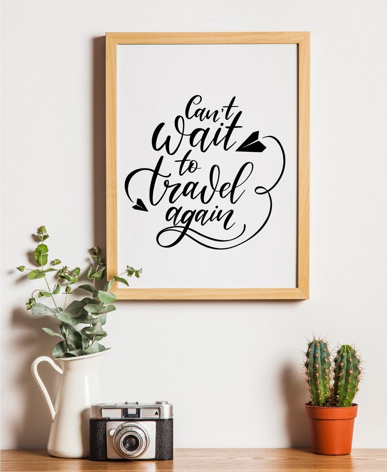 May include: A framed print with the text "Can't wait to travel again" in black calligraphy with a paper airplane graphic. The print is on a white background and is framed in a light wood frame. The print is hanging on a white wall.