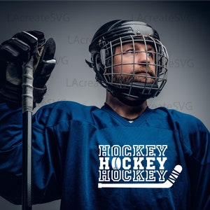 May include: A person in a blue hockey jersey and a black helmet with a cage. The jersey displays the word "HOCKEY" in white, stacked three times, with a hockey puck and stick graphic.