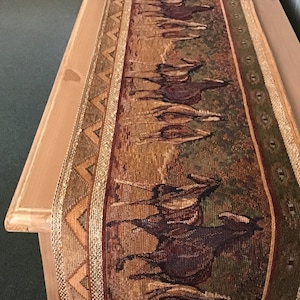 May include: A brown and gold table runner with a woven design of horses running through a field. The runner has a geometric border with gold accents.
