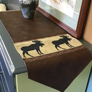 May include: A brown leather table runner with a tan and burgundy patterned border featuring two black moose silhouettes.