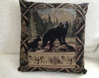 May include: Square decorative pillow with a tapestry design featuring a black bear and cub in a forest setting. The pillow has a black background with a diamond pattern and a border with pine cones and acorns.
