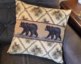May include: A decorative throw pillow with a beige background featuring a repeating pattern of pine branches and pine cones. A brown band across the center of the pillow features two black bears walking through a green forest.