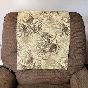May include: A brown recliner chair with a decorative headrest cover. The headrest cover features a beige background with a pattern of pine needles and pine cones in shades of brown and tan. The chair has a textured fabric.