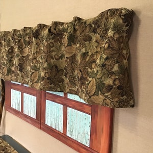 May include: A brown and green valance with a leaf and acorn pattern hangs above a window with a rustic wood frame and a light blue and white patterned glass pane.