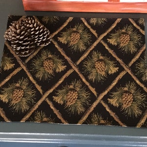 May include: A black placemat with a repeating pattern of pine cones and branches. The placemat has a diamond pattern with brown lines.