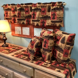 May include: A rustic-themed home decor set featuring a valance, pillows, and a table runner. The fabric has a repeating pattern of deer, trees, and other outdoor elements in shades of red, brown, and tan. The set is displayed on a wooden dresser.