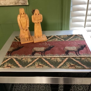 May include: Two carved wooden figures of people stand on a decorative table runner. The runner features a burgundy center with elk silhouettes and green and tan geometric borders. The figures are light brown.