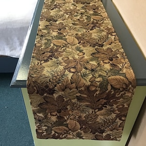 May include: A brown and green table runner with a leaf and acorn pattern. The runner is made of a woven fabric and has a textured surface.