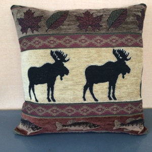May include: A decorative throw pillow with a beige background featuring a repeating pattern of moose silhouettes, fish, and leaves. The pillow has a brown, red, and green striped border.